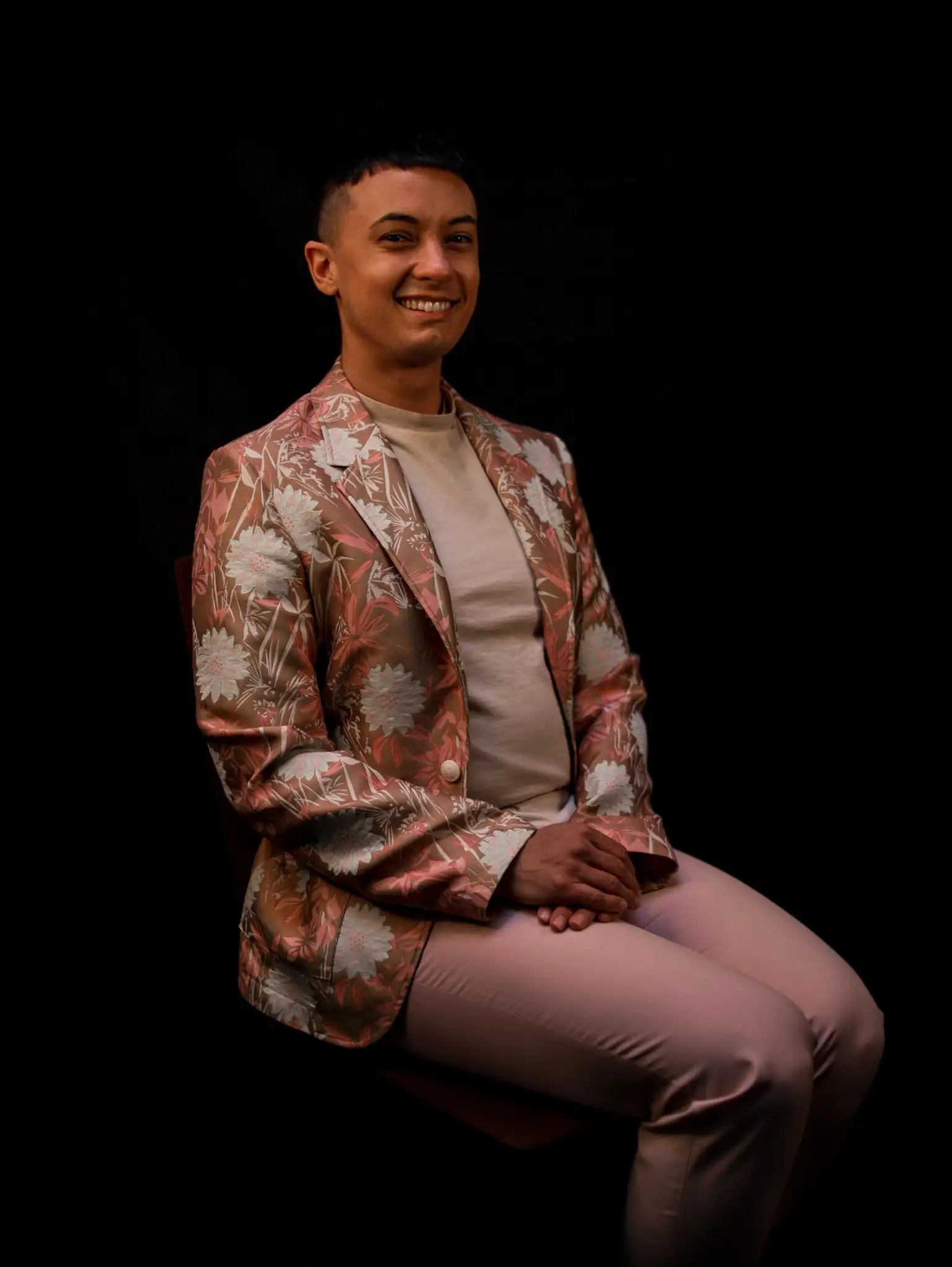 Mel sits in front of a black background in pink pants and a pink and beige floral lamé blazer. They are a white, trans web designer and the owner of The Happy Sites. 